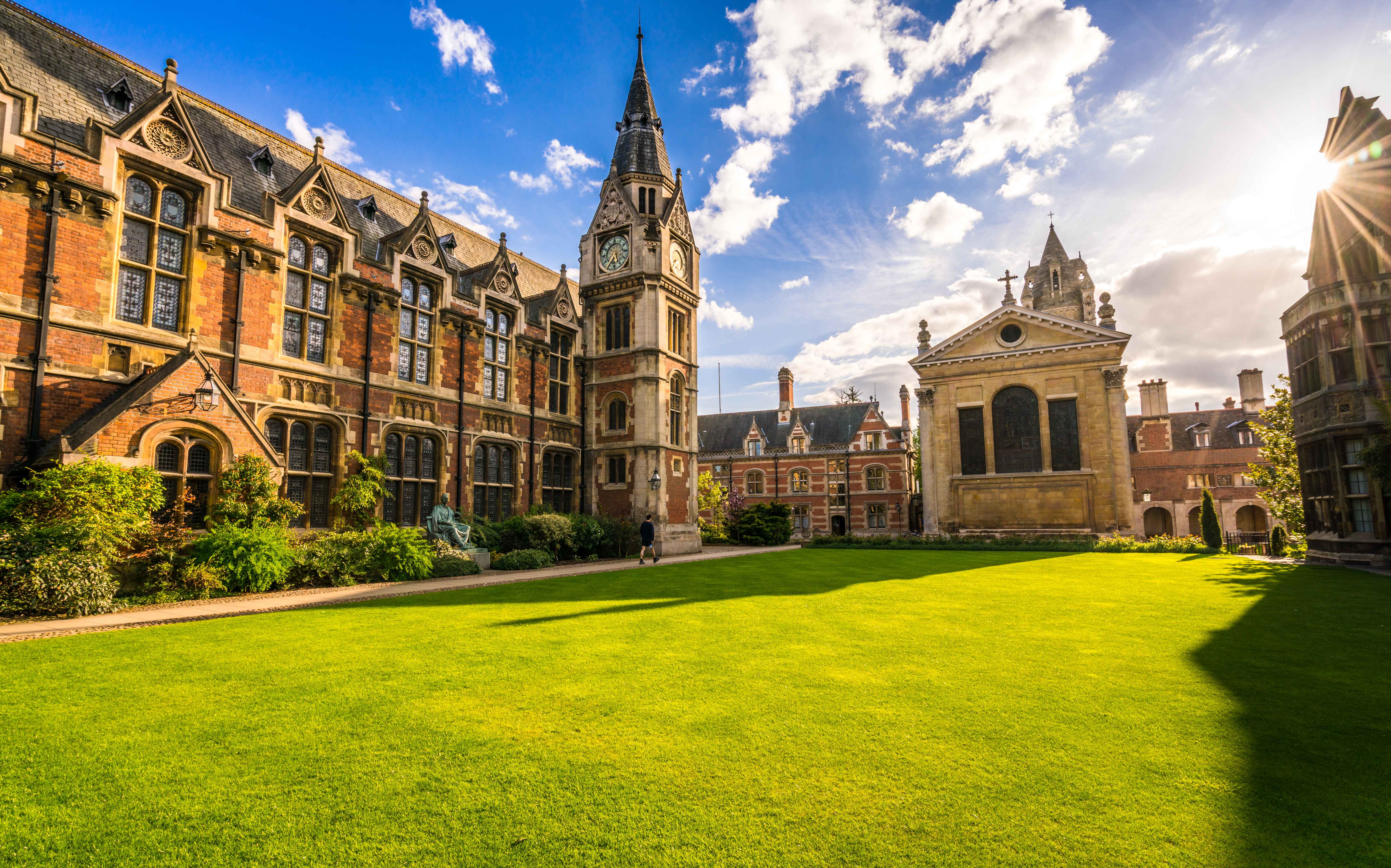 College courtyard with ornate brick buildings, a clock tower, and a chapel, bordered by a green lawn.