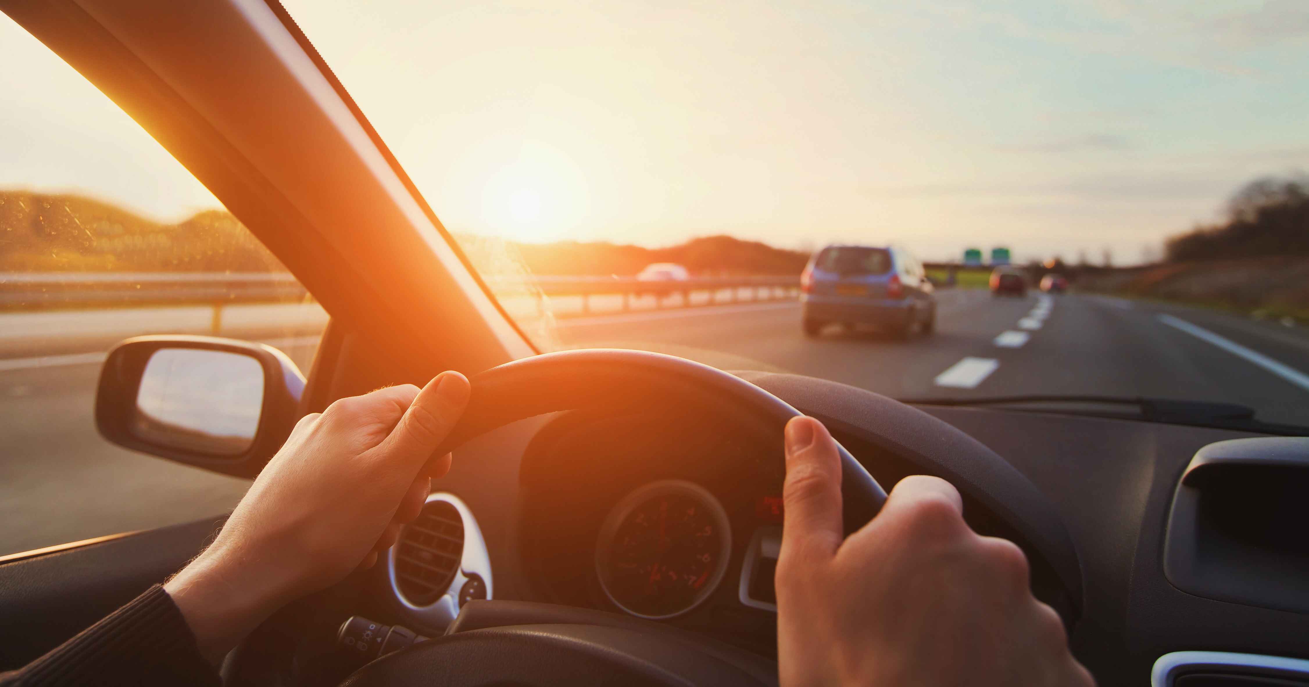 View from inside a car driving on a highway at sunset, with hands on the steering wheel and vehicles ahead.