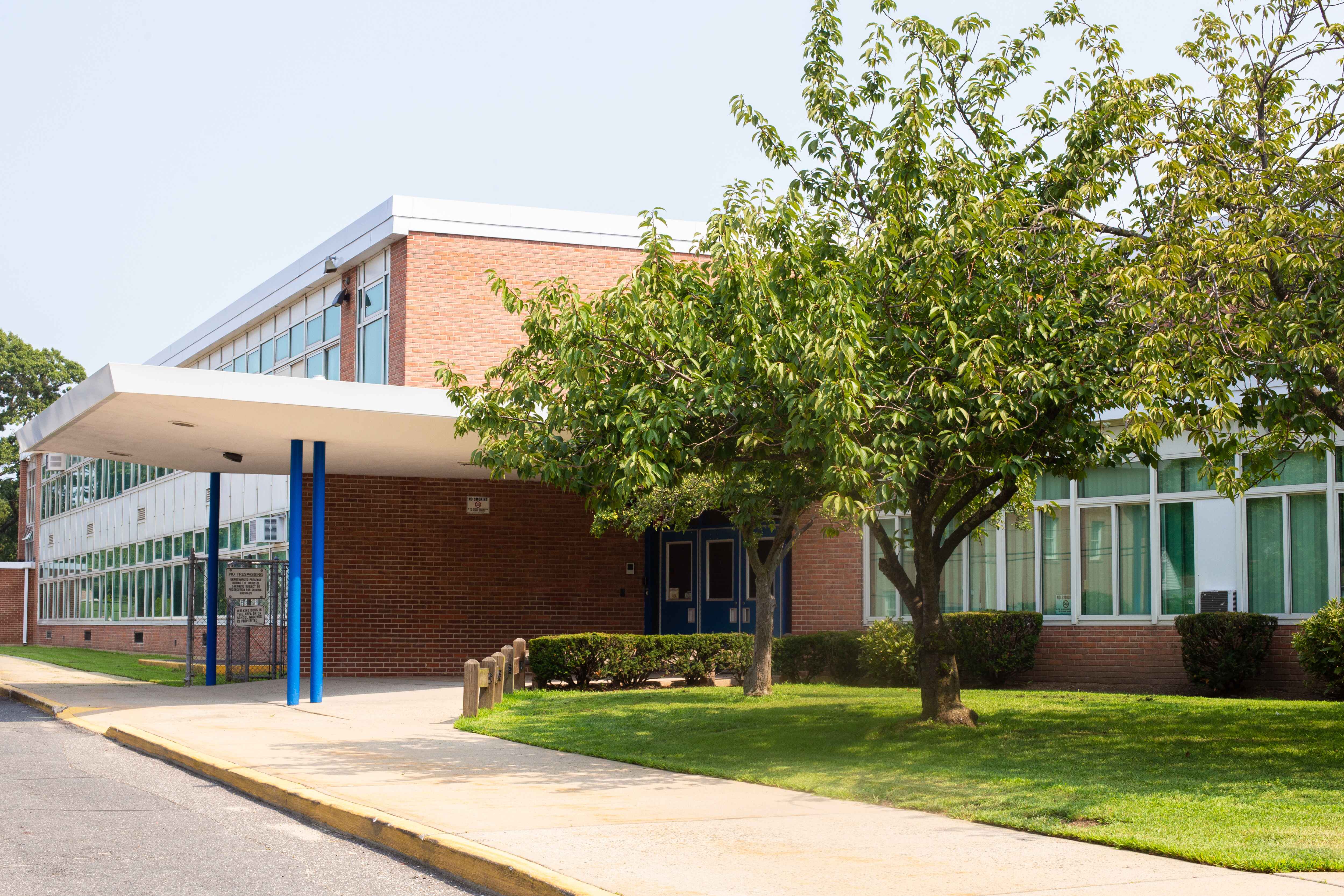 Exterior of a brick school building with large windows, a blue-framed glass entrance, and a flat canopy supported by blue poles