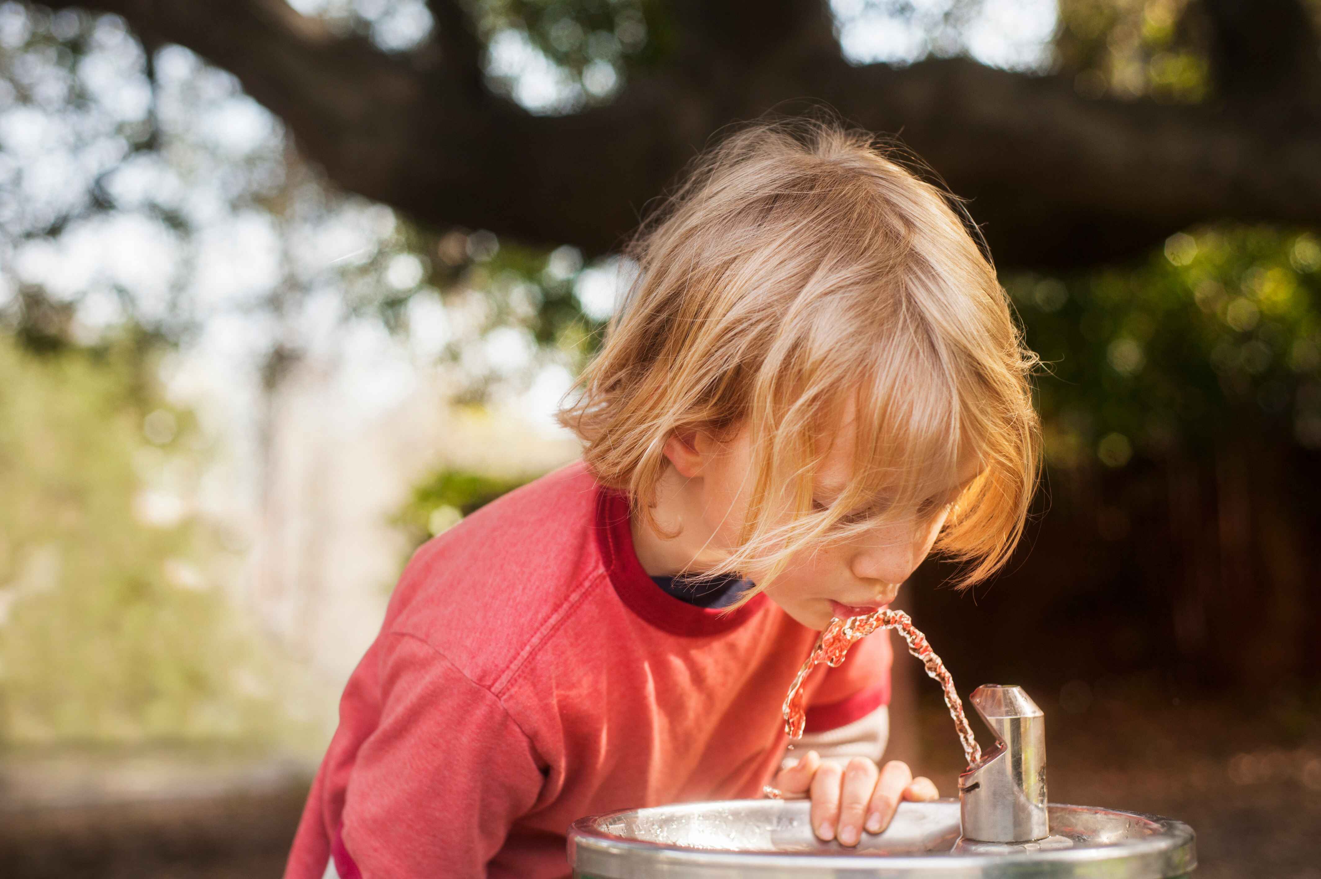 Child drinking from an outdoor water fountain with blurred greenery in the background.