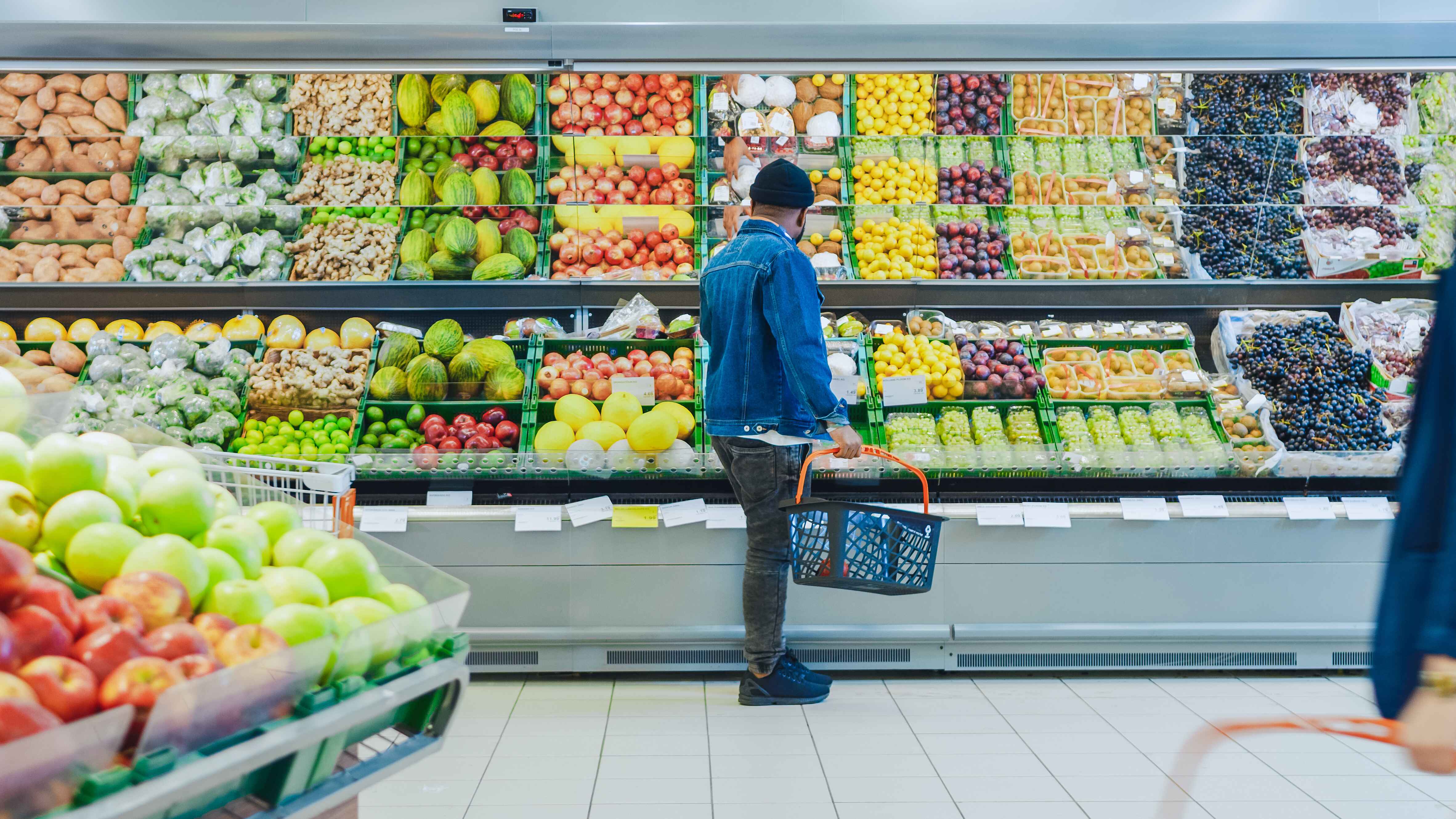 A man with a shopping basket stands in front of a variety of fruits and vegetables in a supermarket.