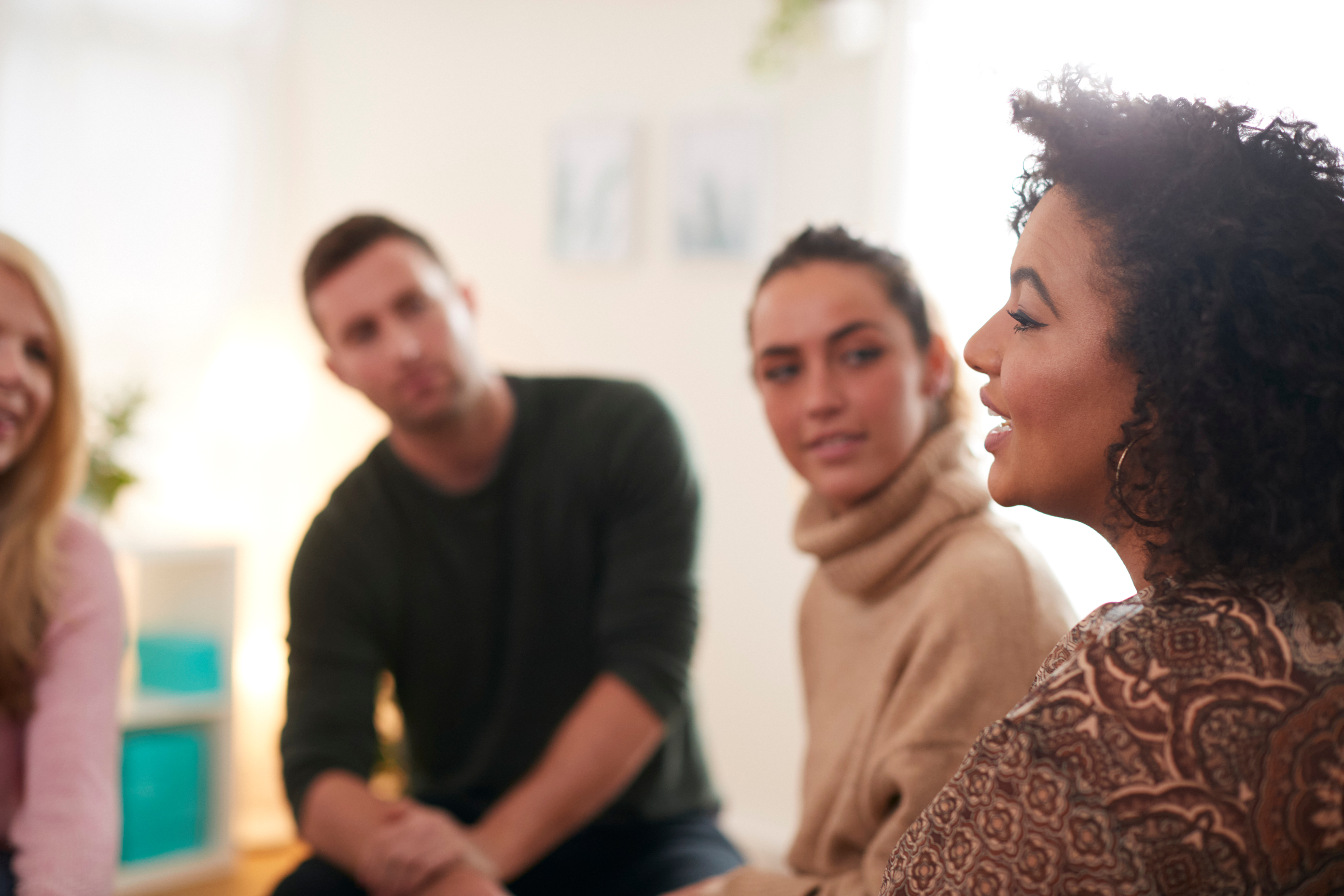 Four people speaking to each other sitting in a circle, a white woman with blonde hair, a white man with brown hair, a white woman with brown hair, and a black woman with curly brown hair