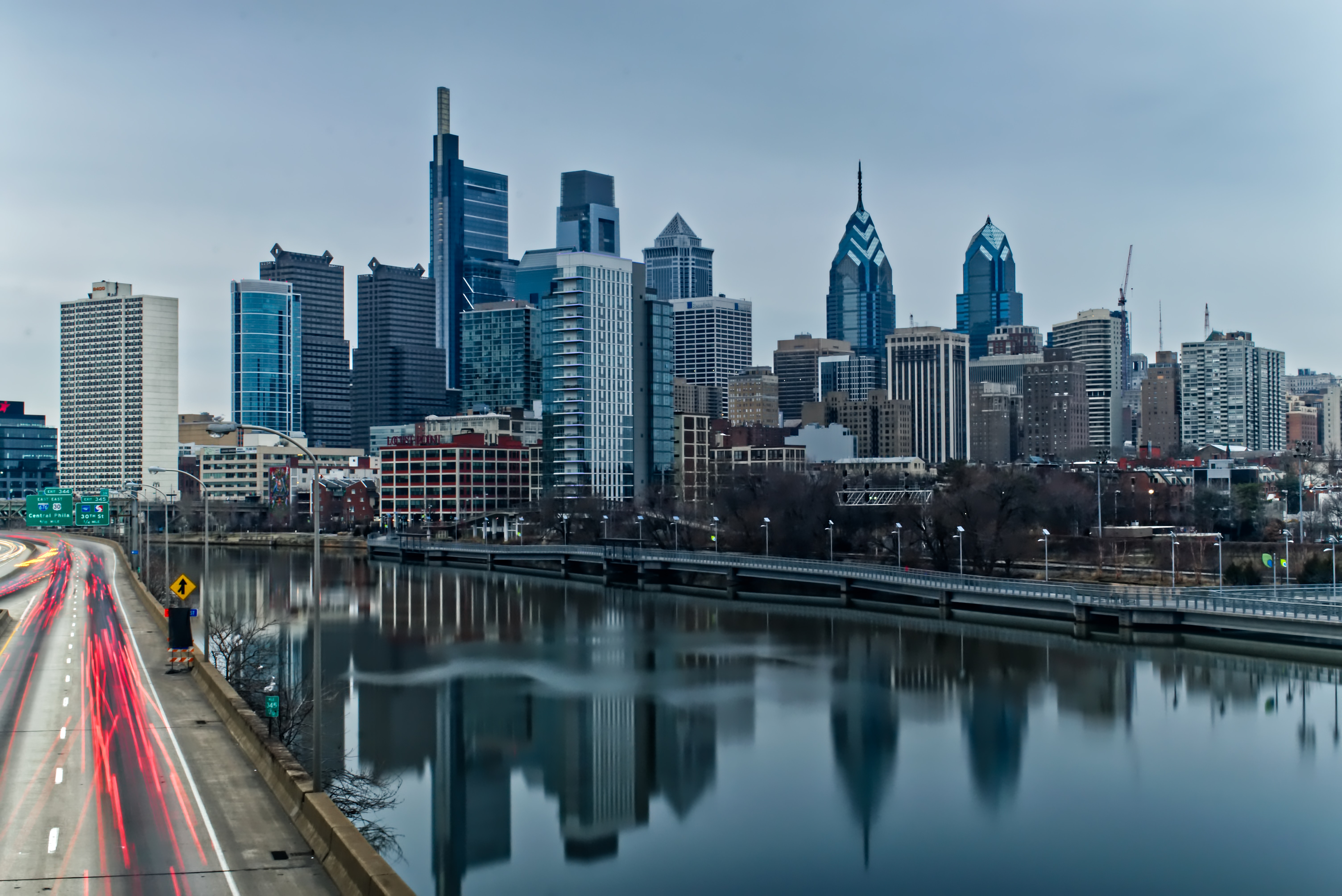 Philadelphia skyline with skyscrapers reflected in a river and highway traffic in the foreground.