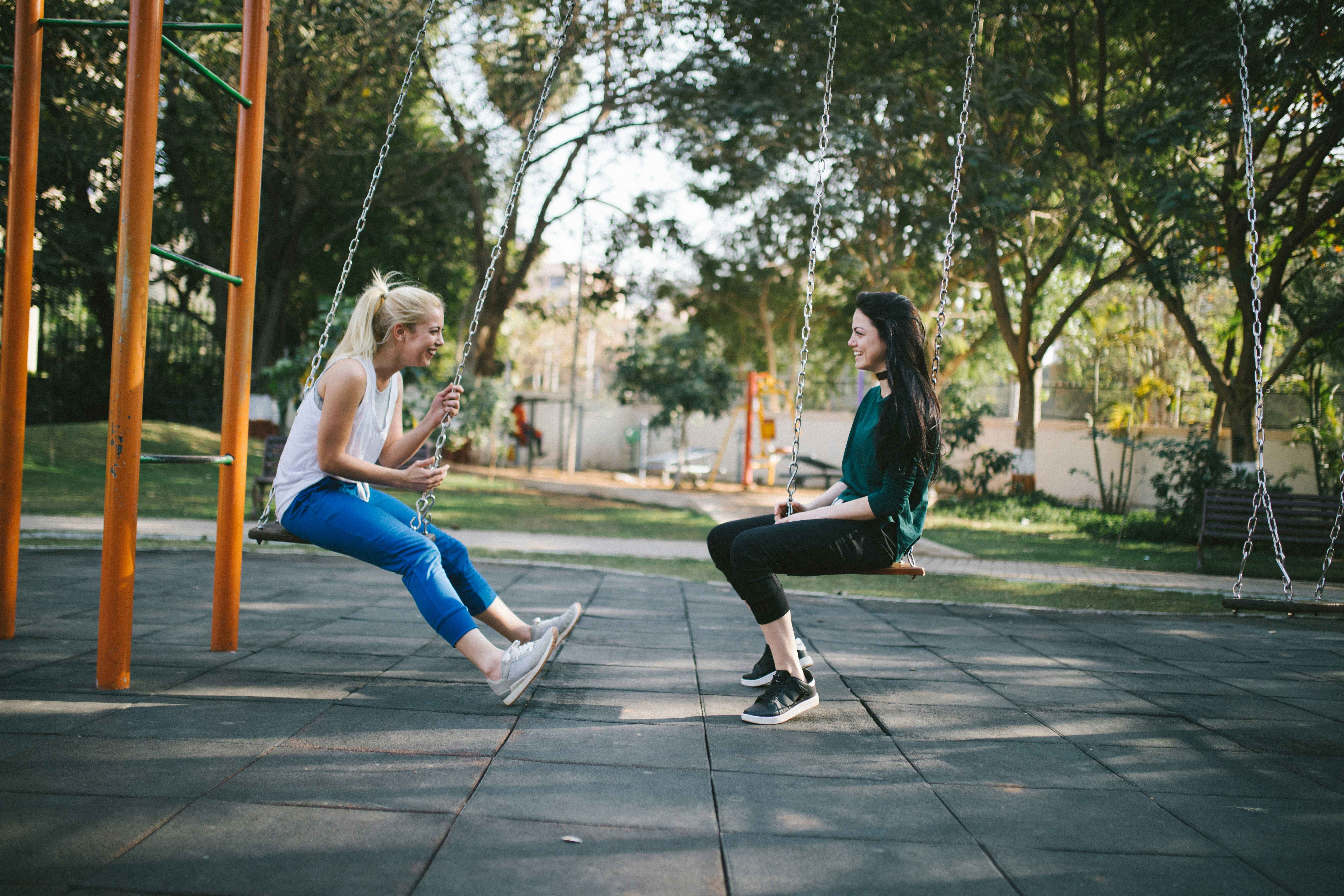 Two people sitting on swings in a park, surrounded by trees.