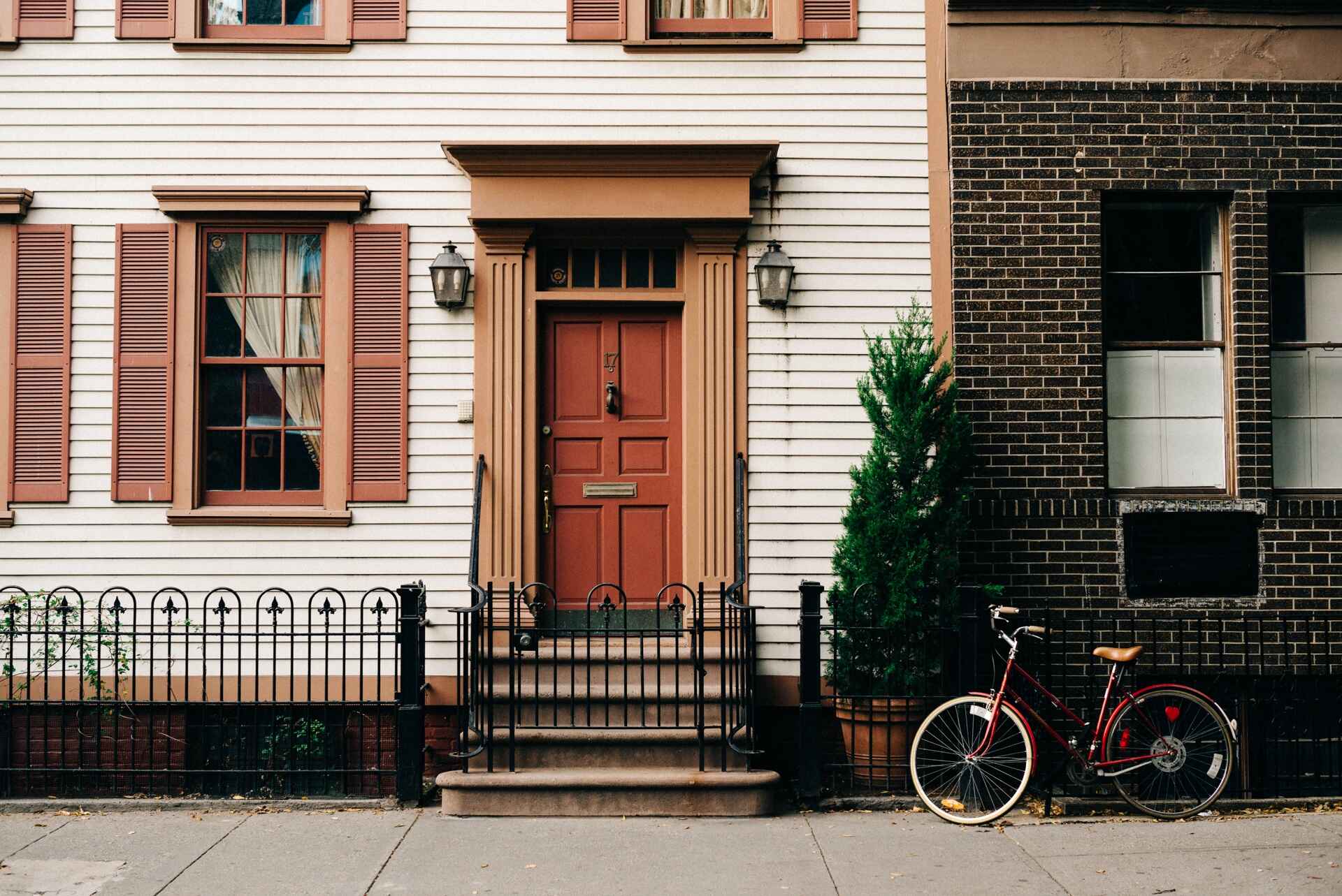 Facade of a traditional house with cream siding, brown shutters, a central entrance, and a red bicycle.