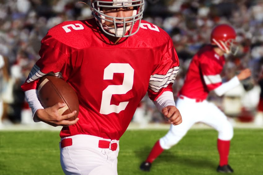 Football player in red jersey holding a football, with another player and a crowd in the background.