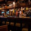 Interior of a bar with shelves of liquor bottles and hanging wine glasses, with a wooden counter and stools.
